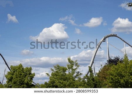 Roller coaster with distinct high arches and tracks set against a clear blue sky and fluffy clouds, surrounded by lush green trees in an amusement park