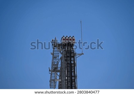A roller coaster train filled with riders sits at the peak of a steep vertical drop, framed against a clear blue sky. The dark metal track and adjacent maintenance staircase emphasize the height, engineering scale, and intense anticipation of the thrill ride.