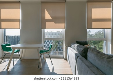 Roller shades automatic on full height windows in the interior. Armchairs with green pillows in the kitchen near windows with motorized roller blinds. Sunny day. Selective fokus. Sunscreen curtains. - Shutterstock ID 2311364883
