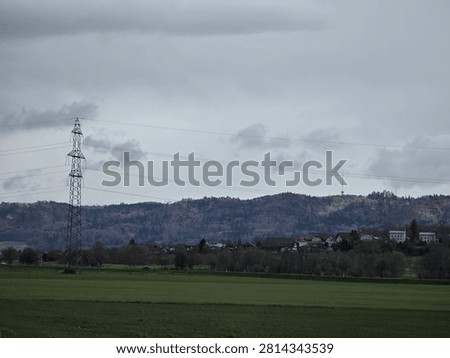 Rolling green fields beneath a dramatic cloudy sky with forested hills, scattered village homes, power lines and transmission tower near Münchenbuchsee, Switzerland, peaceful rural landscape