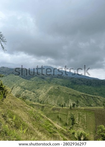 Rolling green hills beneath dramatic dark clouds creating a moody mountain landscape. Natural highland scenery with agricultural fields and untouched environment.