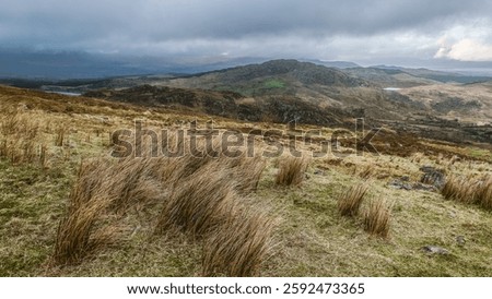 Rolling hills covered with sparse grass under a moody sky create a tranquil atmosphere. Views include distant mountains and a tranquil lake reflecting the surroundings.