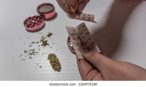 Rolling medical marijuana joint with colored paper in woman hands. Cannabis buds and grinder on white table