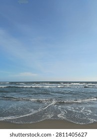 Rolling Waves and Sandy Beach. This dynamic seascape captures multiple rows of gentle, rolling ocean waves breaking near the shore, their crests creating layers of white sea foam that stretch horizontally across the frame. The waves break onto the edge of a dark, wet sandy beach under a light blue sky with faint, streaky clouds.