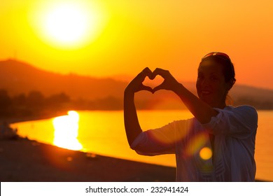 Romance and love at the seaside ; Smiling young woman hands showing symbol of heart on a beautiful beach at sunset