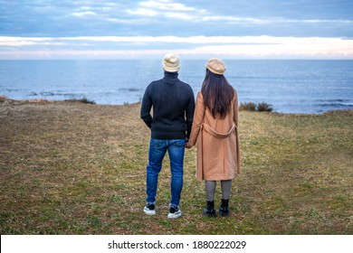 Romance Scene Of Young Heterosexual Couple View From Back Holding Hands Watching The Horizon On The Sea - Two Unrecognizable People Wearing Fall-winter Clothes Looking The Ocean Sunset With Cloudy Sky