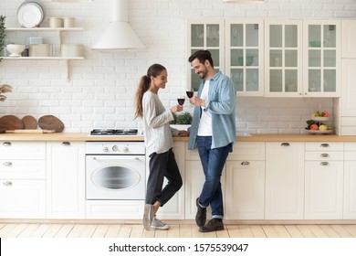 Romantic affectionate beautiful young couple holding glasses standing in modern cozy white kitchen room interior, happy married guy husband and girl wife drinking red wine celebrate together at home