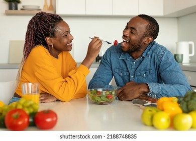 Romantic african american couple eating healthy salad in kitchen, happy wife feeding her husband with tomato, sitting at table and smiling - Powered by Shutterstock