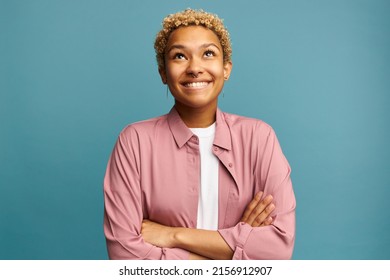 Romantic African American female looking up biting lower lip, making wish, standing over blue background with folded arms, wearing earrings, having nose piercing and blonde short hair, dressed in pink - Shutterstock ID 2156912907