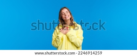 Romantic alluring blond caucasian girlfriend in yellow hoodie, press hands to heart and close eyes with lovely smile recall beautiful memory, imaging perfect date, standing blue background.