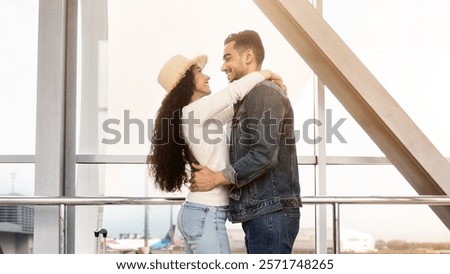 Romantic Arab Couple Hugging While Waiting For Flight In Airport Terminal, Middle Eastern Newlyweds Ready For Honeymoon Travel, Standing With Suitcases Near Window In Lounge Zone, Side View