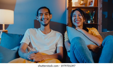 Romantic asia couple man and woman smile and laugh lay down on sofa in living room at night watch comedy movie on television together at home. Married couple family lifestyle, stay at home concept.