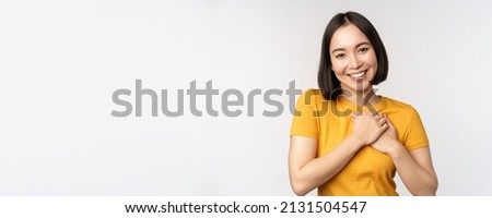 Romantic asian girfriend, holding hands on heart, smiling with care and tenderness, standing in yellow tshirt over white background