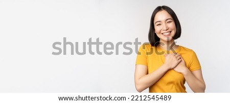 Romantic asian girfriend, holding hands on heart, smiling with care and tenderness, standing in yellow tshirt over white background