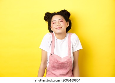 Romantic asian girl with cute pink makeup and summer clothes, close eyes and smiling dreamy, standing on yellow background