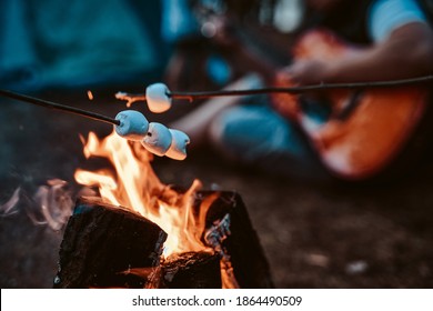 Romantic and atmospheric photography of marshallow on sticks its toasting at the stake in blurred background of guitarist and forest.