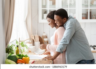 Romantic Black Couple Cleaning Dishes And Bonding In Kitchen, Making Domestic Chores Together. Loving African American Man Cuddling Wife From The Back, Enjoying Spending Time At Home, Side View