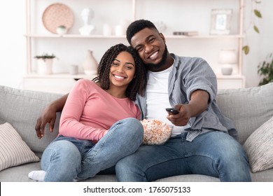 Romantic black couple with popcorn watching tv at home, embracing and smiling at camera