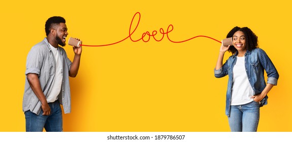 Romantic black guy sending love message through tin phone to his happy african girlfriend, cheerful lovers having fun on valentine's day, standing over yellow background, panorama with copy space