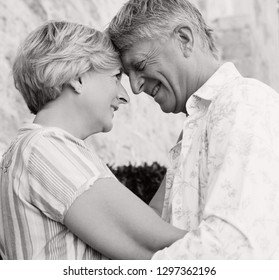 Romantic Black And White Portrait Of Senior Tourist Couple Hugging On Holiday With Heads Together, Smiling Outdoors. Mature Mand And Woman Enjoying Leisure Travel Lifestyle, Retirement Activities. 