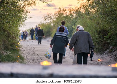 Romantic Bonfire Night At Seaside During Sunset. People Gathering Together To Celebrate Night Of Ancient Lights. Old Couple Walking On Wooden Pathway Towards Sea