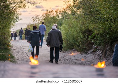 Romantic Bonfire Night At Seaside During Sunset. People Gathering Together To Celebrate Night Of Ancient Lights. Old Couple Walking On Wooden Pathway Towards Sea 