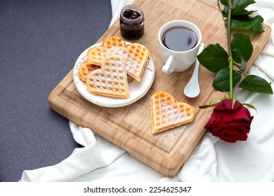 Romantic breakfast in bed on wooden tray with fresh baked homemade heart shaped Belgian waffles, jam, cup of tea or coffee and red rose on gray couch. Valentine's Day breakfast surprise, top view. - Shutterstock ID 2254625347