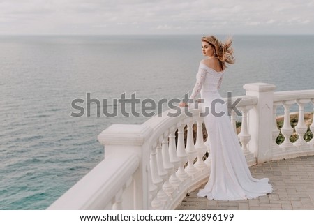 Romantic bride, blond girl in white wedding dress with open shoulders posing on open terrace with backdrop of the sea and rocks. Stylish young woman standing on terrace and looking on ocean at sunset