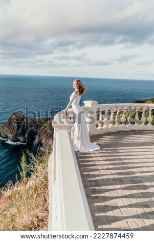 Romantic bride, blond girl in white wedding dress with open shoulders posing on open terrace with backdrop of the sea and rocks. Stylish young woman standing on terrace and looking on ocean at sunset