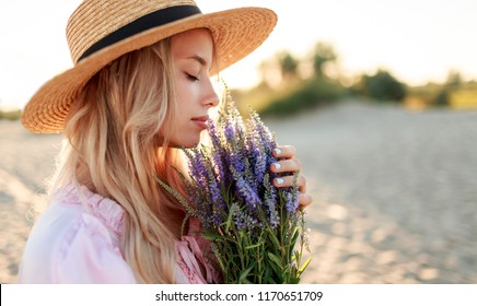  Romantic close up portrait  o charming blonde girl in straw hat  smells    flowers   on   the evening beach,  Warm sunset colors. Bouquet of lavender. 