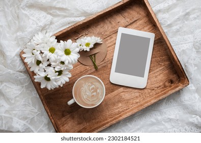 Romantic composition with cup of coffee, white flowers and e-reader with copy space on wooden tray. Template for feminine blog social media.