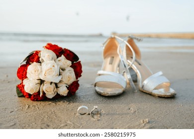  A romantic composition featuring a pair of wedding rings resting on soft golden sand, accompanied by elegant bridal shoes and delicate roses. Perfect for themes related to weddings, engagements.