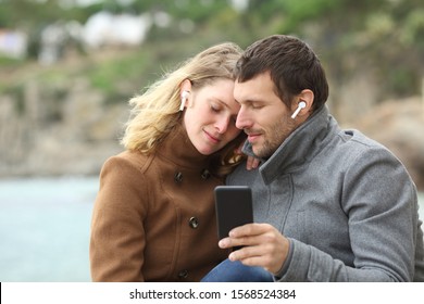 Romantic Couple Of Adults Listening To Music Together Using Mobile Phone And Sharing Earbuds In Winter On The Beach