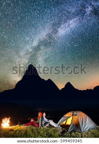 Romantic couple backpackers sitting by bonfire near tent under incredibly beautiful starry sky. Silhouette of the mountains and luminous village in the valley at night in background. Astrophotography