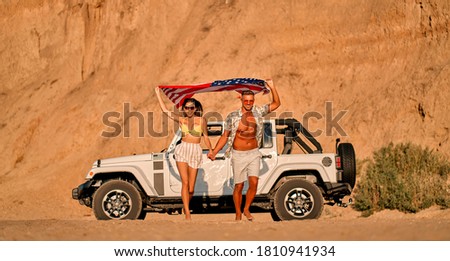 Romantic couple with car parked on the beach. Summer vibes. Traveling together to the sea. Handsome man and beautiful woman walking with American flag in hands