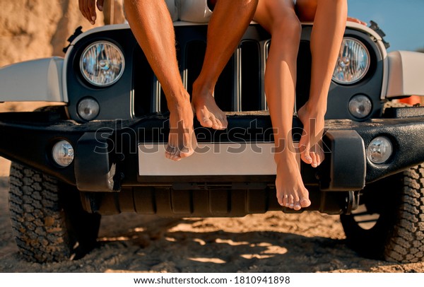 Romantic couple with car parked on the beach. Summer\
vibes. Traveling together to the sea. Cropped image of legs hanging\
down from car.