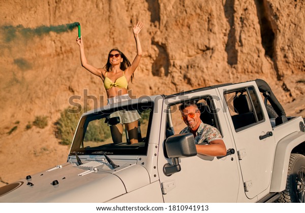 Romantic couple with car\
parked on the beach. Summer vibes. Traveling together to the sea.\
Beautiful woman in swimsuit holding colored smoke while standing in\
car.