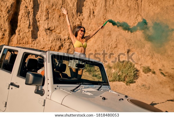 Romantic couple with car\
parked on the beach. Summer vibes. Traveling together to the sea.\
Beautiful woman in swimsuit holding colored smoke while standing in\
car.