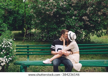 Romantic couple have a date in the city-garden. Young hipsters couple are sitting on the 
bench, kissing and hugging. Wearing casual trendy clothes.