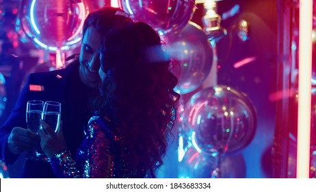 Romantic Couple Holding Champagne Under Confetti In Slow Motion. Affectionate Man And Woman Dancing Slow Dance At Party. Love Couple Hugging On Neon Lamps Background.