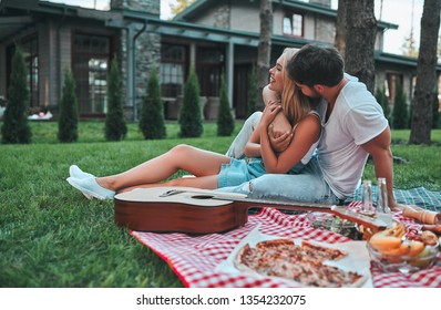 Romantic Couple Hugging While Having Picnic. Handsome Bearded Man And Attractive Young Woman Are Sitting Together On Grass On The Backyard.