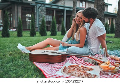 Romantic Couple Hugging While Having Picnic. Handsome Bearded Man And Attractive Young Woman Are Sitting Together On Grass On The Backyard.