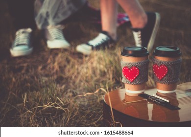 Romantic Couple Hugging While Having Picnic In Sunset Light. Coffee Cup Cozy Knitted Sleeve With Felt Red Heart.