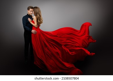 Romantisches Paar in Liebe tanzen. Mann in schwarzem Hemd umarmt geheimnisvolles Mädchen in fliegendem rotem Kleid. Modefrau auf Long Wave Gown und Freund auf grauem Hintergrund
