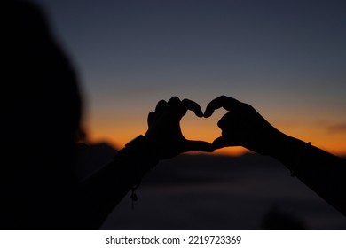 Romantic Couple In Love Gesturing Silhouette Hands Heart Sign, Heart Shape Over Sunslight Sky Background. View Mountaion Light With Mist Morning On Lifestyle Holiday. Focus On Hands Shape