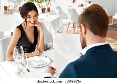 Romantic Couple In Love Having Dinner In Luxury Gourmet Restaurant. Happy Beautiful Lovely People Reading Menu, Choosing Food, Celebrating Anniversary Or Valentine's Day. Romance And Relationships.