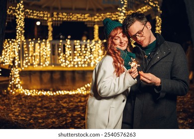 A romantic couple in love holding sparklers outdoors and celebrating christmas and new year's eve. A jolly couple is hugging on city street downtown illuminated by christmas lights and decorations. - Powered by Shutterstock