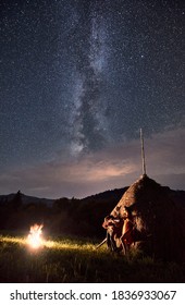 Romantic couple near haystack basks by the fire under starry sky on which Milky Way is visible. Guy with guitar in his hands is singing for girl with valley of hills at background
