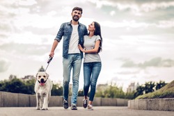 Romantic Couple Is On A Walk In The City With Their Dog Labrador. Beautiful Young Woman And Handsome Man Are Having Fun Outdoors With Golden Retriever Labrador.