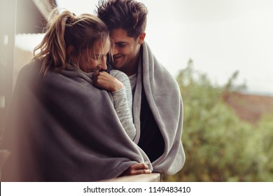 Romantic couple on a winter holiday. Man and woman standing together in a hotel room balcony wrapped in blanket. Couple embracing and smiling. - Powered by Shutterstock